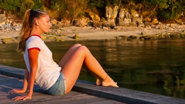 Young woman sttiing on the pier by the sea at sunset background — Stock Video