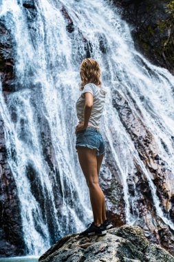 Güneş batarken kumlu sahilde şapkalı ve sarı elbiseli beyaz bir kadın batan güneşe bakıyor.