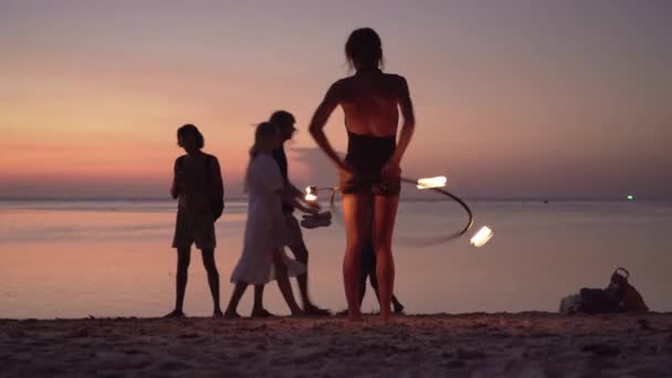 夕暮れ時に浜辺で火をつけてフラフープで踊る若い女性のシルエット 人々はビーチを歩き カラフルな夕日を楽しむ — ストック動画