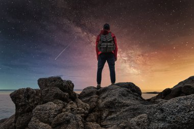 Sırtında sırt çantası olan kırmızı ceketli bir adam gece deniz kenarındaki kayalıklarda duruyor ve yıldızlı gökyüzüne ve denizin üzerindeki Samanyolu 'na bakıyor.