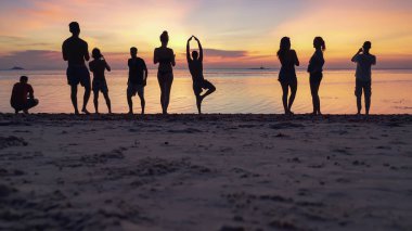 Kumlu sahilde gün batımının keyfini çıkaran siluetler ve denizin arka planında akıllı telefonlardan fotoğraf çeken insanlar. 
