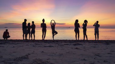 Kumlu sahilde gün batımının keyfini çıkaran siluetler ve denizin arka planında akıllı telefonlardan fotoğraf çeken insanlar. 