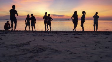 Kumlu sahilde gün batımının keyfini çıkaran siluetler ve denizin arka planında akıllı telefonlardan fotoğraf çeken insanlar.