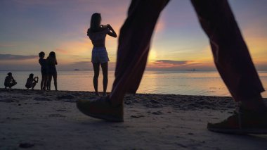 Kumlu sahilde gün batımının keyfini çıkaran siluetler ve denizin arka planında akıllı telefonlardan fotoğraf çeken insanlar. 