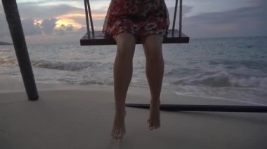 Gün batımında sahilde sallanan salıncakta sallanan elbiseli mutlu genç bir kadın.