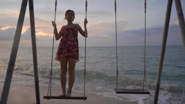 日落时 身穿连衣裙的年轻快乐女子在海滩上荡秋千 站在水面上 — 图库视频影像