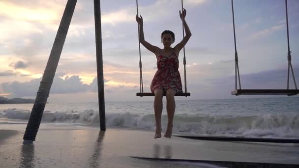 日落时 身穿连衣裙的年轻快乐女子在海滩上荡秋千 站在水面上 — 图库视频影像