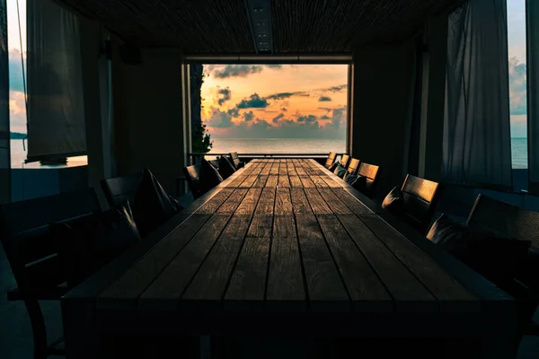 Room on the beach with a long wooden table with chairs against the backdrop of an open window at a spectacular colorful sunrise over the sea