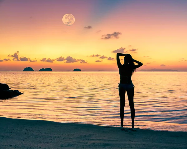 在夏日的傍晚 一个美丽的日落和一轮巨大的满月映衬下 一位年轻女子在海边的轮廓 — 图库照片