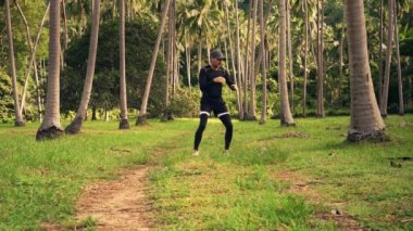 Siyah antreman kıyafetleri içinde atletik beyaz bir adam ve bir şapka güneşli bir yaz sabahı çimenli bir palmiye çayırında kendini savunmak için gölgeli boks eğitimi veriyor. Yavaş çekim videosu.