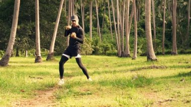 Siyah antreman kıyafetleri içinde atletik beyaz bir adam ve bir şapka güneşli bir yaz sabahı çimenli bir palmiye çayırında kendini savunmak için gölgeli boks eğitimi veriyor. Yavaş çekim videosu.