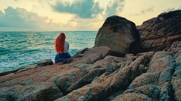 Young woman alone with long pink hair sits on the rock and looks at the sea at sunrise