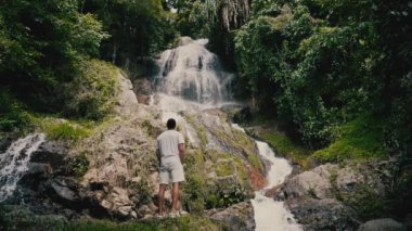 Beyaz giysili genç bir adam kayalara tırmanıyor ve güneşli bir yaz gününde tropiklerde bir şelaleye bakıyor.