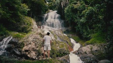 Beyaz giysili genç bir adam kayalara tırmanıyor ve güneşli bir yaz gününde tropiklerde bir şelaleye bakıyor.