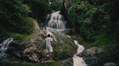 Beyaz giysili genç bir adam kayalara tırmanıyor ve güneşli bir yaz gününde tropiklerde bir şelaleye bakıyor.