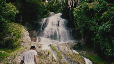 Beyaz giysili genç bir adam kayalara tırmanıyor ve güneşli bir yaz gününde tropiklerde bir şelaleye bakıyor.