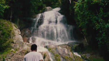 Beyaz giysili genç bir adam kayalara tırmanıyor ve güneşli bir yaz gününde tropiklerde bir şelaleye bakıyor.