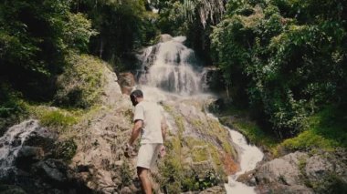 Beyaz giysili genç bir adam kayalara tırmanıyor ve güneşli bir yaz gününde tropiklerde bir şelaleye bakıyor.
