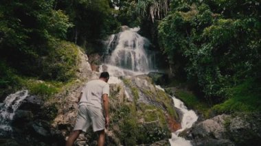 Beyaz giysili genç bir adam kayalara tırmanıyor ve güneşli bir yaz gününde tropiklerde bir şelaleye bakıyor.