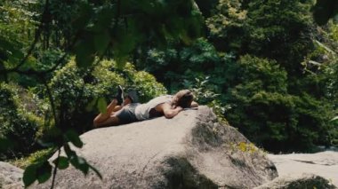 Güneşli bir yaz gününde, tropiklerdeki bir şelalenin yakınındaki bir kayada uzanan spor giysili beyaz kadın akıllı telefon kullanır.