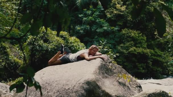 夏の晴れた日に熱帯の滝の近くの岩の上に横たわるスポーツウェアの若い幸せな白人女性とスマートフォンを使用しています — ストック動画