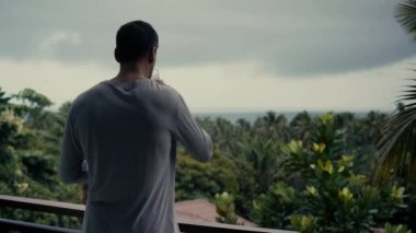 Genç atletik adam evin balkonunda duruyor, denizin üzerinde dramatik yağmur bulutları gibi görünüyor ve akıllı telefondan bir film çekiyor.