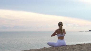 Genç kadın yogası rengarenk günbatımında deniz kıyısında kumda oturuyor.
