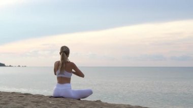 Genç kadın yogası rengarenk günbatımında deniz kıyısında kumda oturuyor.