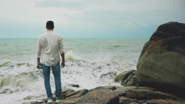 Beyaz gömlekli ve kot pantolonlu genç cesur bir adam fırtına sırasında bir kayanın üzerinde duruyor ve denize bakıyor.