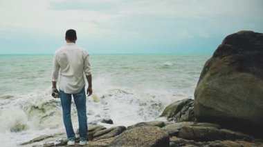 Beyaz gömlekli ve kot pantolonlu genç cesur bir adam fırtına sırasında bir kayanın üzerinde duruyor ve denize bakıyor.