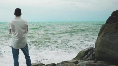Beyaz gömlekli ve kot pantolonlu genç cesur bir adam fırtına sırasında bir kayanın üzerinde duruyor ve denize bakıyor.