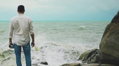 Beyaz gömlekli ve kot pantolonlu genç cesur bir adam fırtına sırasında bir kayanın üzerinde duruyor ve denize bakıyor.