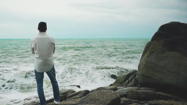 一个身穿白衬衫和牛仔裤的勇敢的年轻人 在暴风雨中站在岩石上凝视着大海 — 图库视频影像