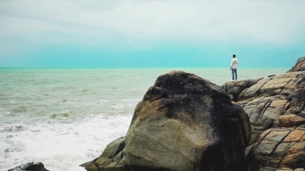 嵐の中岩の上に立って海を見ている白いシャツとジーンズの若い勇敢な男 — ストック動画