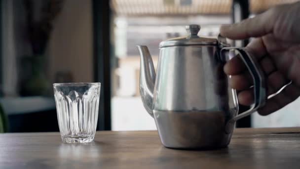 在街上咖啡店的一张木制桌子上 一只用传统的铁制茶壶把茶倒入一个透明的杯子中的男性亲密手 — 图库视频影像