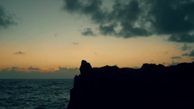 Gün doğumunda deniz kenarındaki kayalıklarda duran bir adamın silueti.