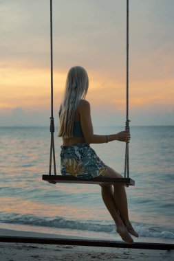 Sarışın, uzun saçlı, kumlu bir sahilde sallanan ve günbatımının tadını çıkaran beyaz bir kadın.