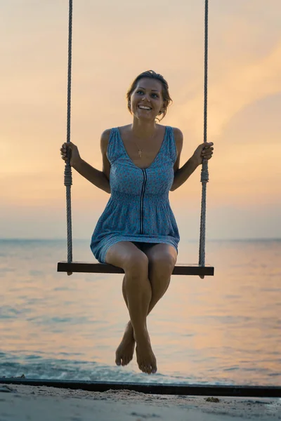 Young happy smiling caucasian woman in dress swinging on a swing standing by the sea on the  beach at sunset