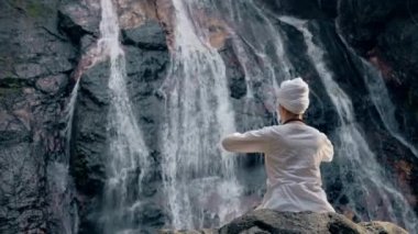 Beyaz elbiseli genç bir kadın bir kayanın üzerinde şelalenin arka planında oturuyor ve yoga yapıyor.