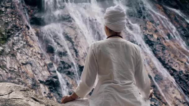 白い服の若い大人の女性は滝の背景に岩の上に座ってヨガを練習 — ストック動画