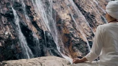 Beyaz elbiseli genç bir kadın bir kayanın üzerinde şelalenin arka planında oturuyor ve yoga yapıyor.