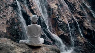 Beyaz elbiseli genç bir kadın bir kayanın üzerinde şelalenin arka planında oturuyor ve yoga yapıyor.