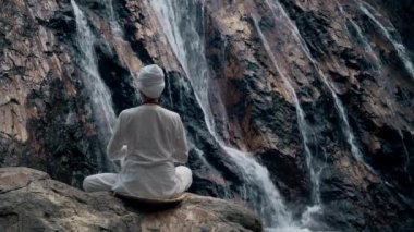 Beyaz elbiseli genç bir kadın bir kayanın üzerinde şelalenin arka planında oturuyor ve yoga yapıyor.