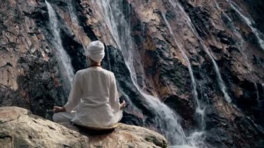 Beyaz elbiseli genç bir kadın bir kayanın üzerinde şelalenin arka planında oturuyor ve yoga yapıyor.