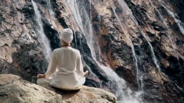 Beyaz elbiseli genç bir kadın bir kayanın üzerinde şelalenin arka planında oturuyor ve yoga yapıyor.