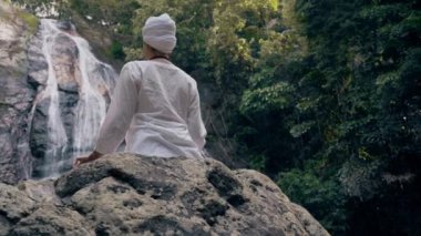 Beyaz elbiseli genç bir kadın bir kayanın üzerinde şelalenin arka planında oturuyor ve yoga yapıyor.