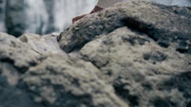 Beyaz elbiseli genç bir kadın bir kayanın üzerinde şelalenin arka planında oturuyor ve yoga yapıyor.