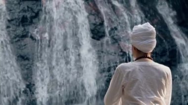 Beyaz elbiseli genç bir kadın bir kayanın üzerinde şelalenin arka planında oturuyor ve yoga yapıyor.