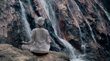 Beyaz elbiseli genç bir kadın bir kayanın üzerinde şelalenin arka planında oturuyor ve yoga yapıyor.