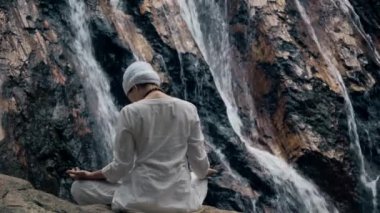 Beyaz elbiseli genç bir kadın bir kayanın üzerinde şelalenin arka planında oturuyor ve yoga yapıyor.